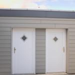 Zwei weiße Türen mit Diamantfenstern an einem Holzhaus mit grauer Fassade.