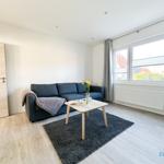 Wohnzimmer mit Sofa, Tisch und Fenster. Holzboden und graue Gardinen.