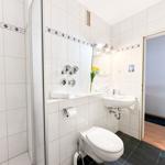 Badezimmer mit Dusche, Waschbecken und Toilette. Weiße Fliesen und schwarze Bodenfliesen.