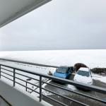 Blick vom Balkon auf schneebedecktes Gelände mit zwei parkenden Autos.