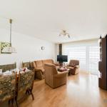 Wohn- und Essbereich mit Sofa, Sesseln und Esstisch. Holzboden und Fenster mit Vorhängen.