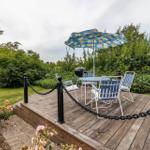 Terrasse mit Tisch, Stühlen, Grill und Sonnenschirm. Umgeben von Grün und Blumen.
