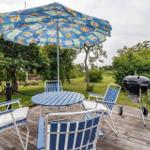 Terrasse mit Tisch, Stühlen, Sonnenschirm und Grill. Umgeben von Grün und Bäumen.