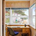 Desk with chair by window, view of wooden houses and trees.