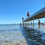 A pier extends into clear water, people enjoy swimming and sunbathing.