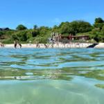 Clear water beach with pier and swimmers under blue sky.