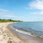 Beach with sand and stones, waves gently break. Trees and houses are visible in the background.