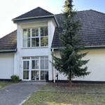 Haus mit weißer Fassade, dunklem Dach und Glasfront. Vorgarten mit Baum und Kiesweg.