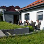 Grüner Garten mit Terrasse und Grill. Gäste sitzen an Tisch. Haus mit rotem Dach.
