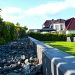 Garten mit Rasenfläche, Steinschüttung und Steinmauer. Im Hintergrund Haus mit rotem Dach.