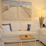 Wohnzimmer mit Sofa, Sessel und Tisch. Wandbild zeigt Strand. Stehlampe mit Holzgestell.