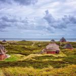Dorfhäuser mit Strohdächern liegen auf Dünen am Meer unter bewölktem Himmel.