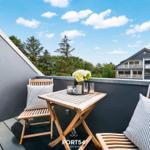 Terrasse mit Holztisch, zwei Stühlen und Blick auf grüne Bäume und ein Haus.