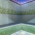 Raum mit grünen Mosaikfliesen, sternenhimmelartigem Deckenlicht und Steinball auf Sockel.