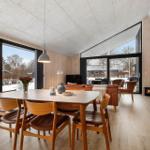 Essbereich mit Tisch und Stühlen, großflächige Fenster, Holzwände und Sitzbereich im Hintergrund.
