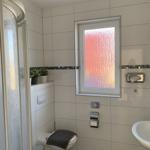 Badezimmer mit Dusche, Toilette und Waschbecken. Fenster mit getöntem Glas.