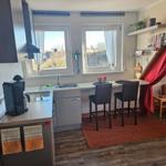 Küche mit Arbeitsplatte, zwei Fenstern und Barhocker. Holzboden und roter Teppich.