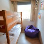 Zimmer mit Holzbett, Fenster und lila Sitzkissen.