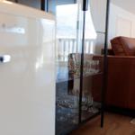 Weißer Kühlschrank neben Glasregal mit Gläsern und Sofa im Hintergrund.
