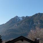 Schneebedeckte Berge mit Wald und Hausdach im Vordergrund.