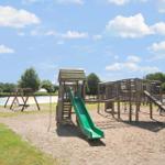 Large playground with slide, climbing frame, and swing on sandy ground under blue sky.