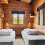 Two beds in a wooden house room with windows and wall shelves.