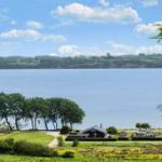 House with view of calm lake and green hills under blue sky.