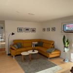 Wohnzimmer mit gelbem Sofa, Tischen und Pflanze. Durchgang zur Küche.