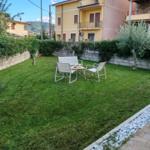 Green garden with seating area and stone wall. Background: yellow house with balcony.