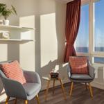 Zwei graue Sessel mit rosa Kissen stehen vor einem Fenster mit Meerblick.