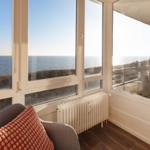 Ein Raum mit Blick aufs Meer, große Fenster, Sitzgelegenheit und Balkon mit Tisch und Stühlen.