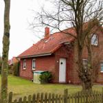 Rotes Ziegelhaus mit weißen Fenstern und Tür. Vorgarten mit Holzzaun und Bäumen.