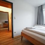 Schlafzimmer mit Bett, Fenster und Holzboden. Durchgang zu Wohnbereich mit TV.