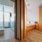 Schlafzimmer mit Holzboden und Schlafplatz. Nebenraum ist die Badewanne mit Dusche.