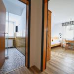 Zimmer mit Holzboden, Bett, Tisch und Stühlen. Nebenraum mit Dusche und Fliesen.