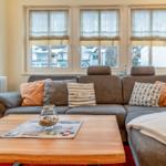 Ein Wohnzimmer mit grauem Sofa, Holztisch und Fenstern mit Rollo.