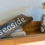 Holzschild mit Aufschrift seaside und Glasflasche mit Muscheln auf Holzregal.