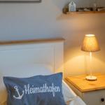 Schlafzimmer mit Bett, Nachttisch und Lampe. Kissen mit Aufschrift Heimathafen.