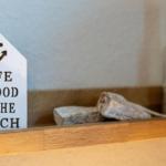 Ein Schild mit Anker und Text steht auf einem Holzregal neben Steinen und einer Flasche.