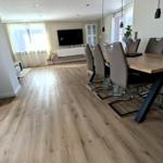 Essbereich mit Holztisch und Stühlen. Im Hintergrund Wohnzimmer mit Fernseher und Fenstern.