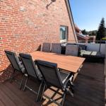Terrasse mit Holztisch, Stühlen und Sitzgruppe. Hintergrund: Hauswand und Nachbarhäuser.