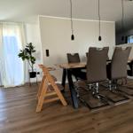 Esszimmer mit Holztisch, braunen Stühlen und Kinderschreibtisch. Durchgang zur Küche und Balkon mit Vorhängen.