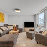 Wohnzimmer mit Sofa, Sessel und Fernseher. Große Fenster mit Blick auf schneebedeckte Bäume.