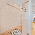Duschkabine mit Mosaikboden und Handbrause. Im Hintergrund ist ein Wohnzimmer mit Sessel sichtbar.