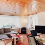 Wohnzimmer mit Sofa, Esstisch und Fernseher. Holzdecke und Fenster mit Vorhängen.