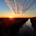 Sonnenuntergang über Fluss und Felder mit spiegelndem Himmel.