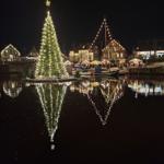 Weihnachtsbaum auf Wasser, beleuchtet, mit Reflexion. Hintergrund: beleuchtete Häuser und Marktstände.