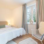 Schlafzimmer mit Bett, Sessel und Fenster mit Blick auf Außenbereich.