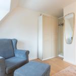 Ein Raum mit grauem Sessel, Kleiderschrank und Spiegel an der Wand.