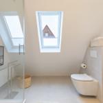 Badezimmer mit Dusche, WC und Fenster. Holzregal und Korb sind sichtbar.